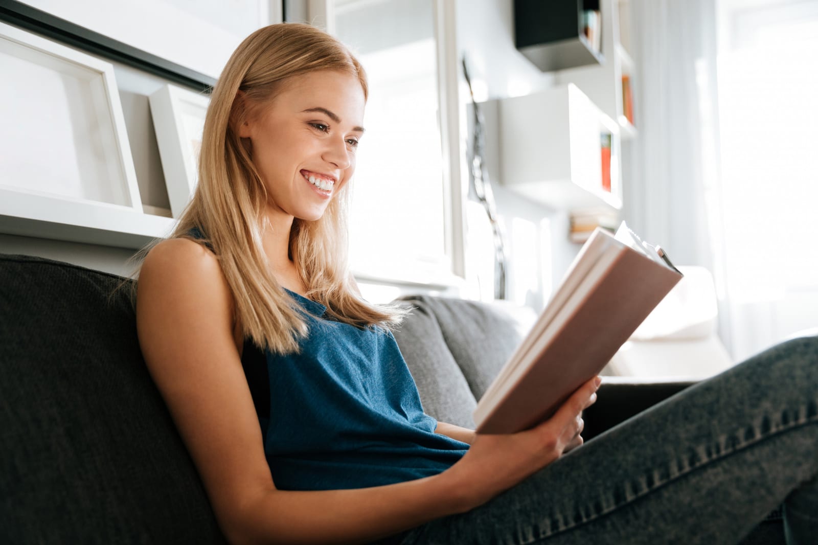 Mulher sorridente sentada no sofá lendo um livro de capa rosa.