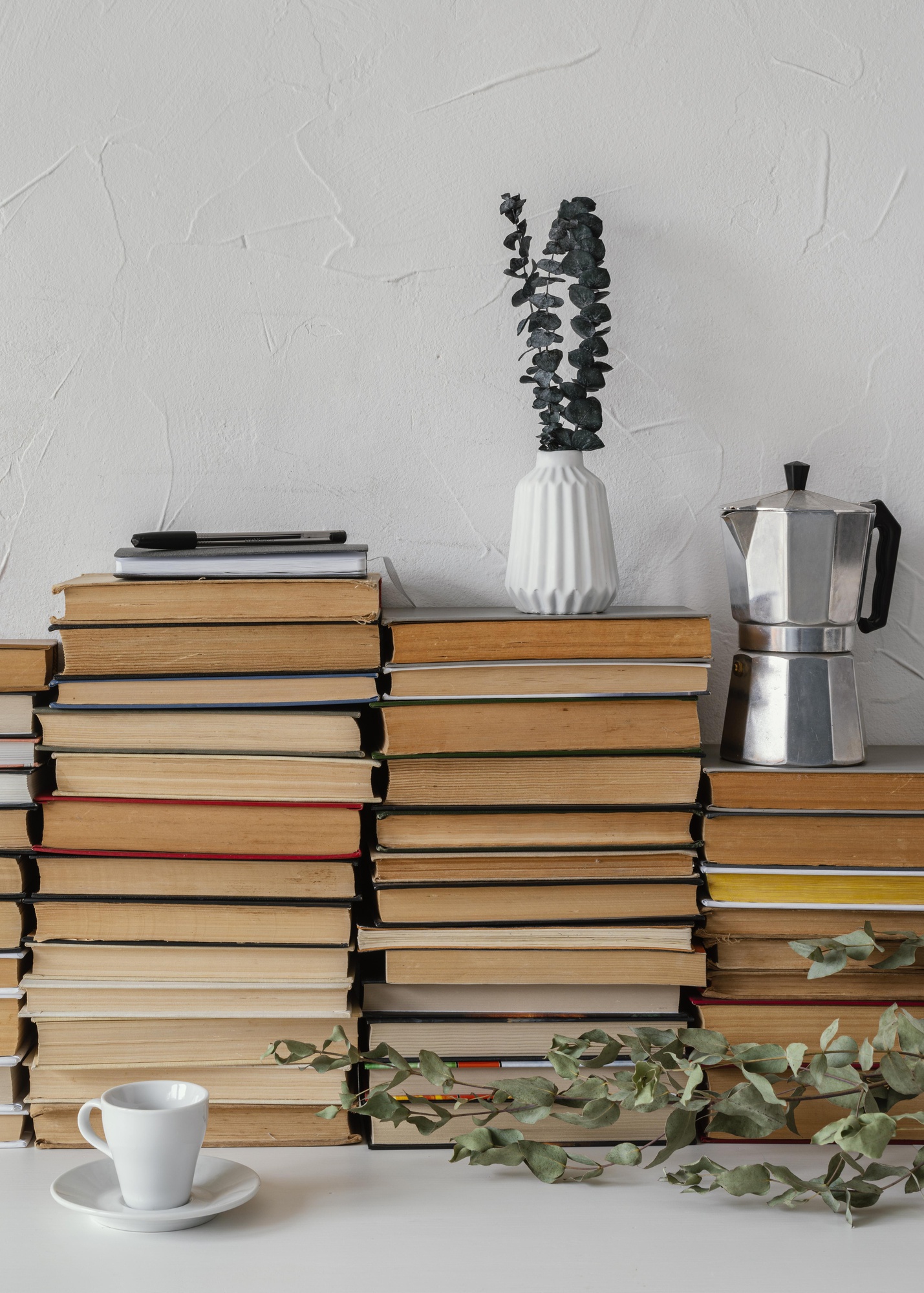 Pilhas de livros sobre uma mesa, com uma cafeteira, uma xícara, uma planta e um caderno com caneta.