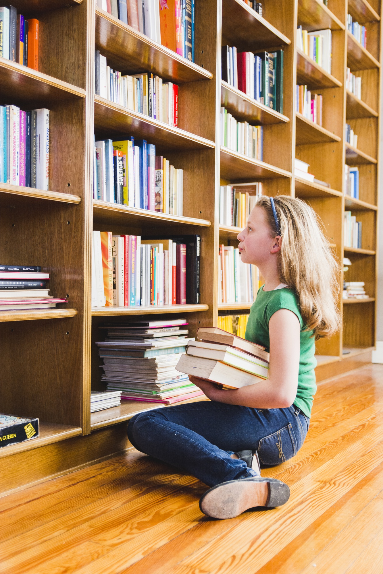 Menina sentada no chão de uma biblioteca doméstica, segurando uma pilha de livros e olhando para a estante.
