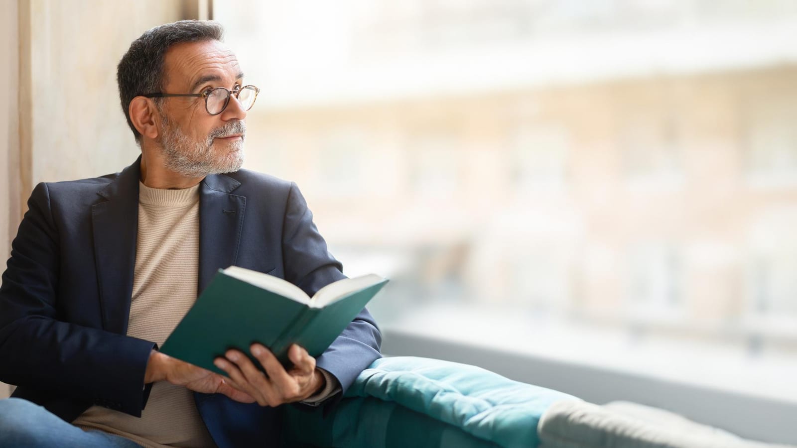 Homem de meia-idade sentado perto de uma janela, lendo um livro de capa dura verde.